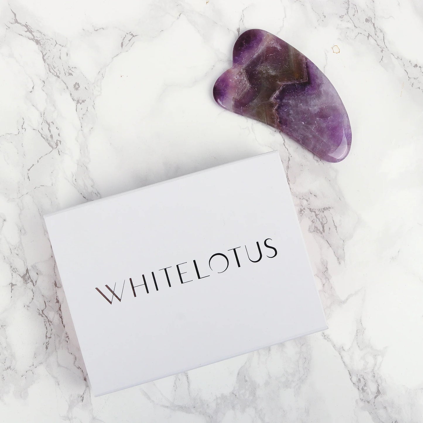 Purple gua sha tool on a white card with 'WHITELOTUS' branding on a marble surface